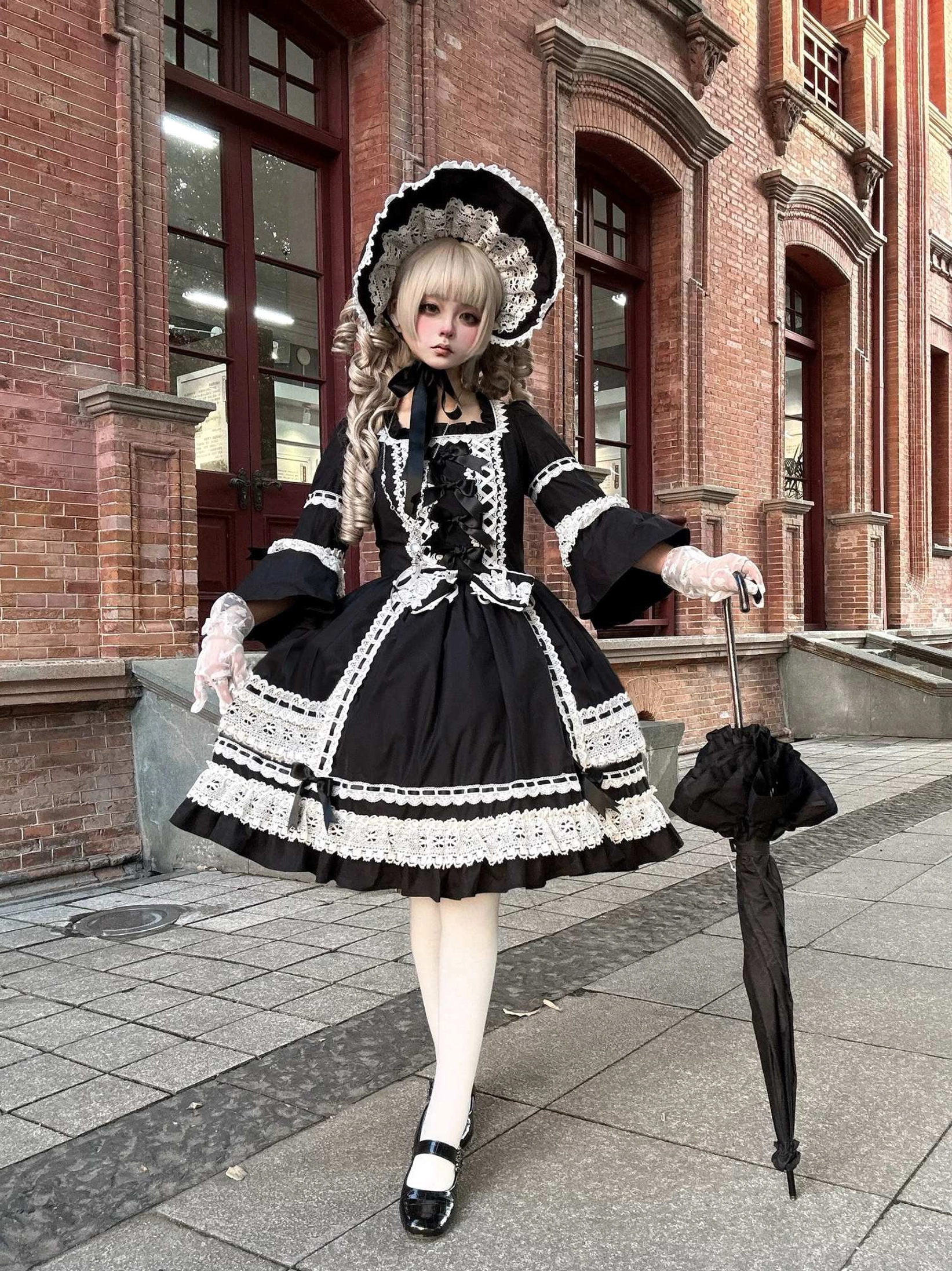 Black old school lolita one-piece dress with white lace front, puff sleeves, and matching bonnet worn outdoors on red brick street with black lace umbrella and white stockings
