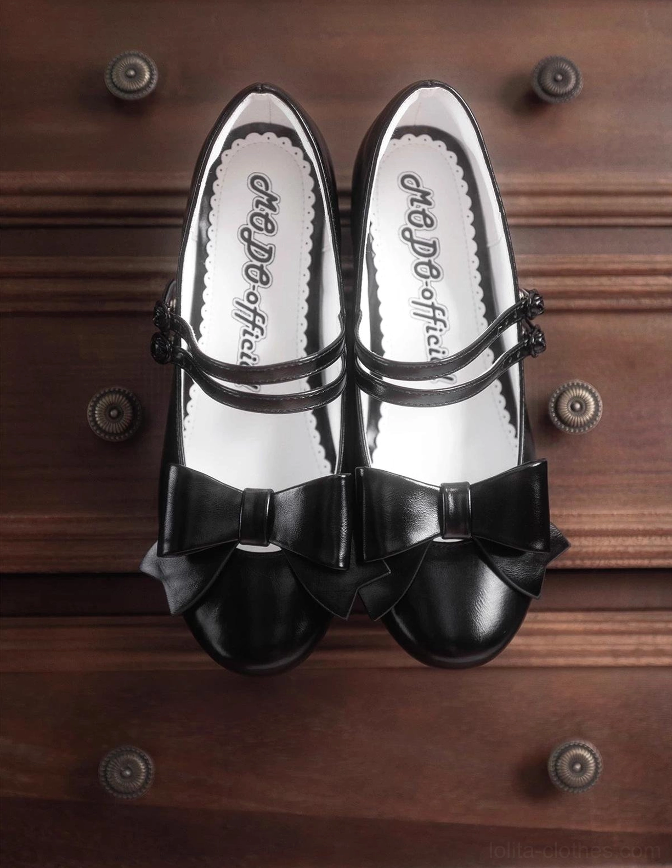 Black pointed toe Lolita Mary Jane shoes with double ankle straps and decorative front bow displayed on vintage wooden dresser showing printed branded text insole detail