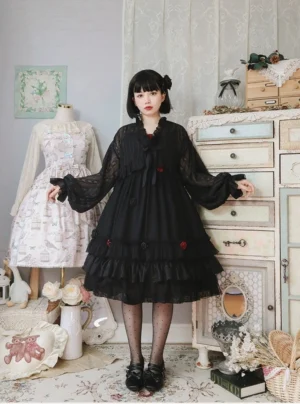Black sheer spring Lolita JSK dress with polka dot chiffon fabric, ruffled neckline with bow detail, layered ruffled hem, and red rose embellishments worn by model in vintage bedroom, front full-body view