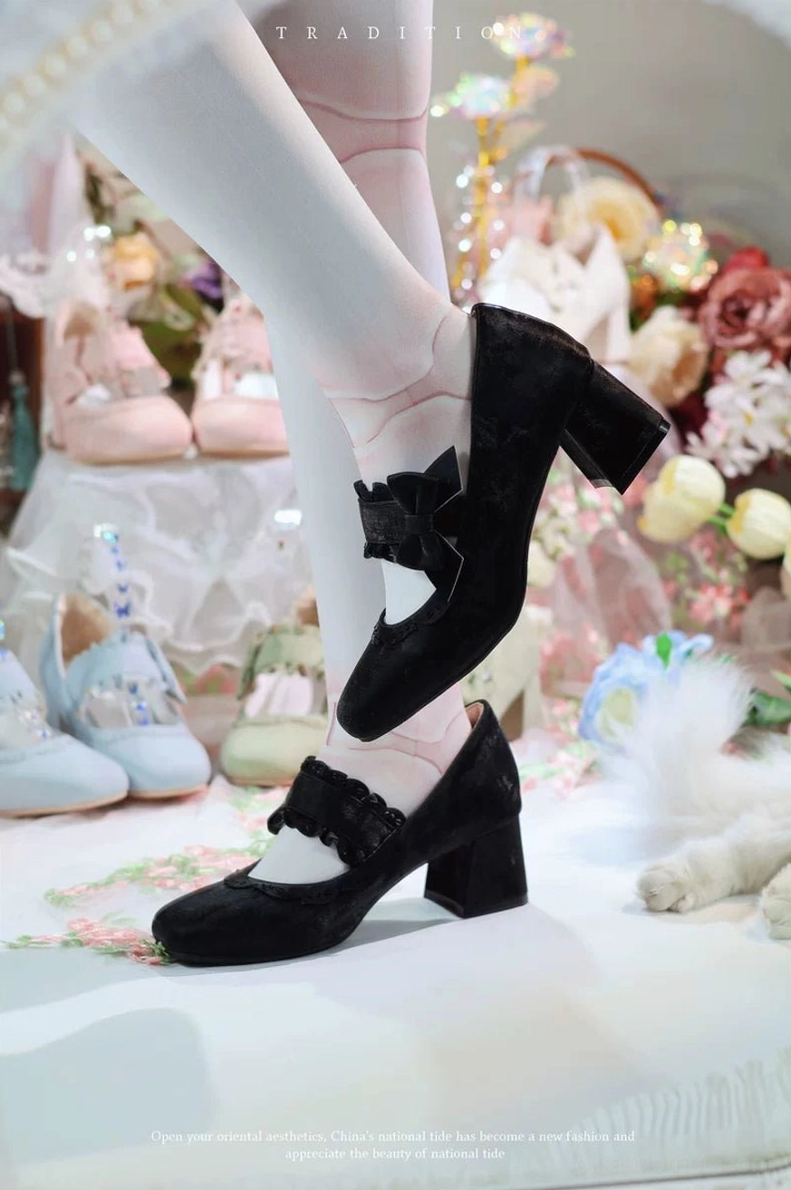 Black suede classic lolita chunky heel mary jane shoes with large decorative bow on ankle strap and matte finish, side view of on-foot wear with pastel lolita shoes and floral bouquet background