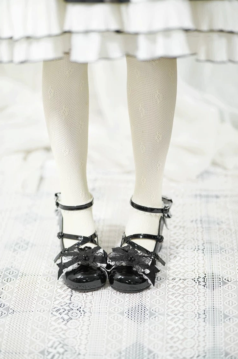 Black glossy patent leather sweet Lolita high heels worn on model's feet in tiered ruffled skirt and sheer lace stockings on lace floor mat, small bow details on cross ankle straps