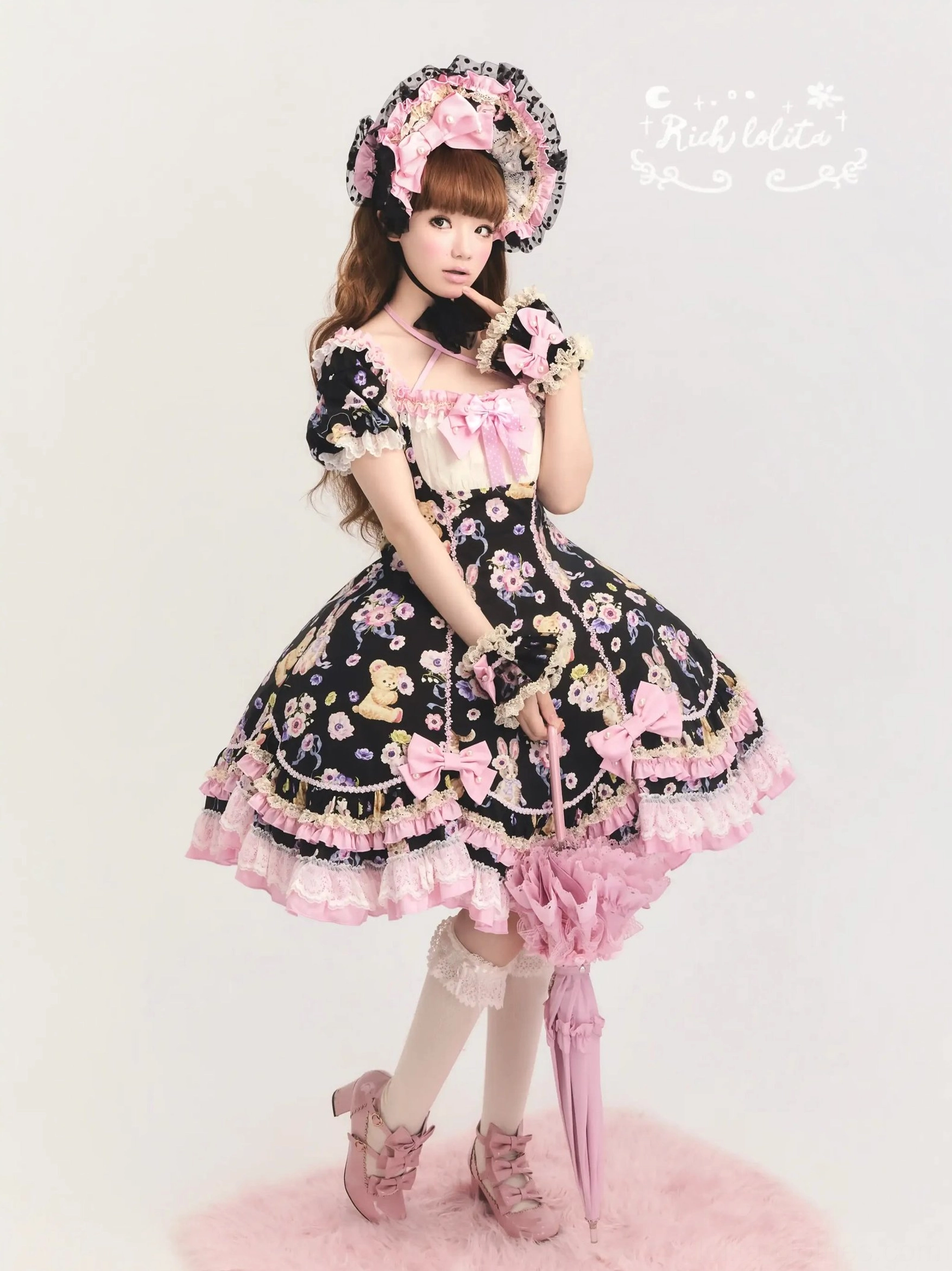 Full-body front view of black old school lolita OP dress with teddy bear floral prints, bow decorations, matching bonnet and gloves, pink lace parasol, studio pink fluffy rug setting