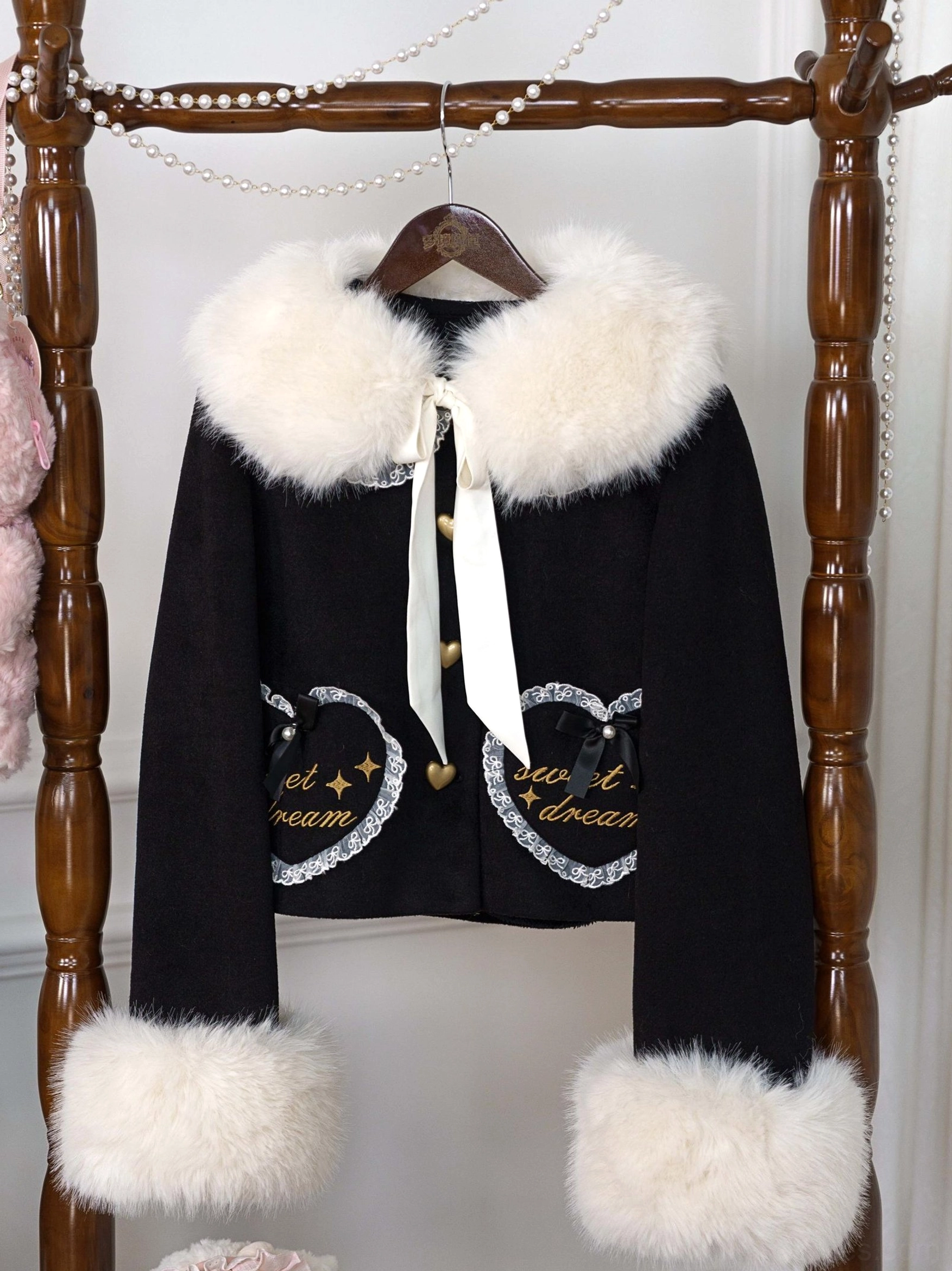 Black fleece-lined sweet lolita coat with lace-trimmed heart embroidery pockets and fluffy faux fur trims, featuring elegant satin neck decoration on wooden rack