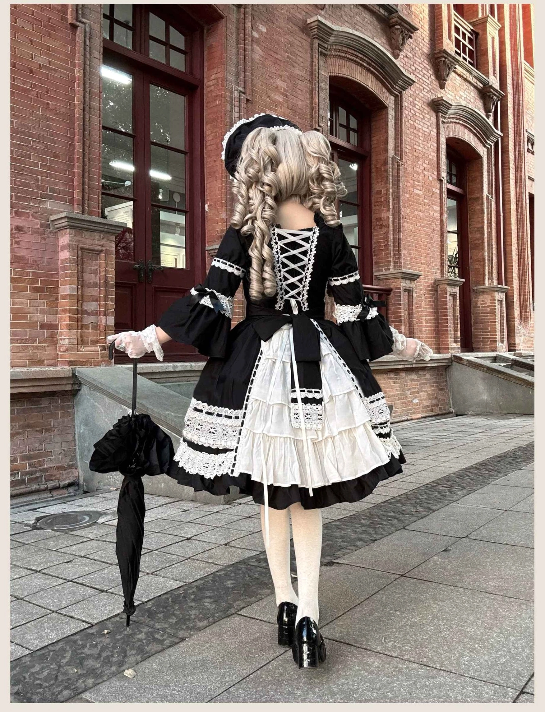 Back view of black lolita OP dress showing lace-up closure, layered white ruffled underskirt, and double-flounce hem detail with matching bonnet and black lace umbrella