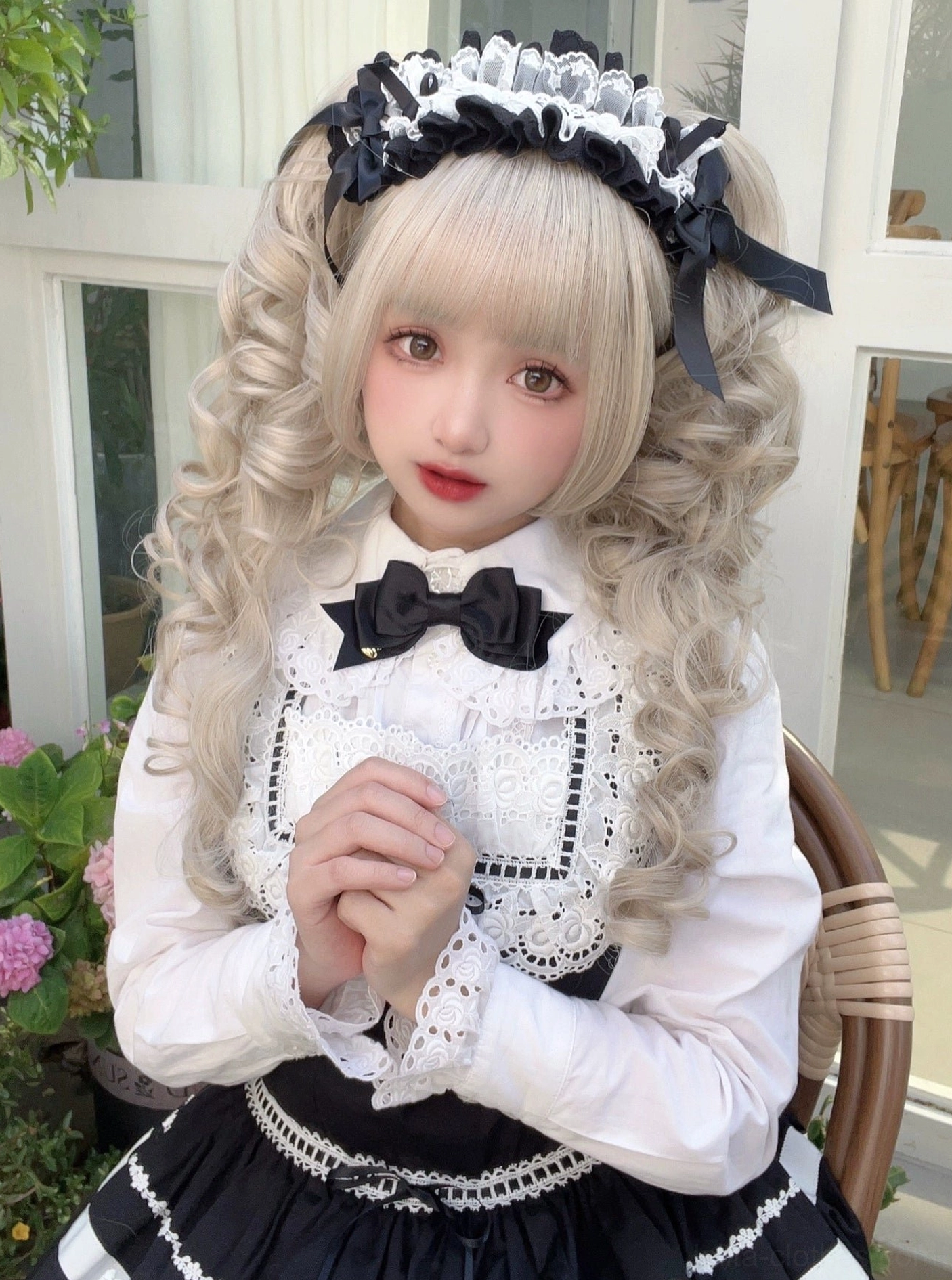 Black and white Lolita maid ruffled lace headband with bow details worn by blonde model in close-up front view indoor balcony garden foliage background