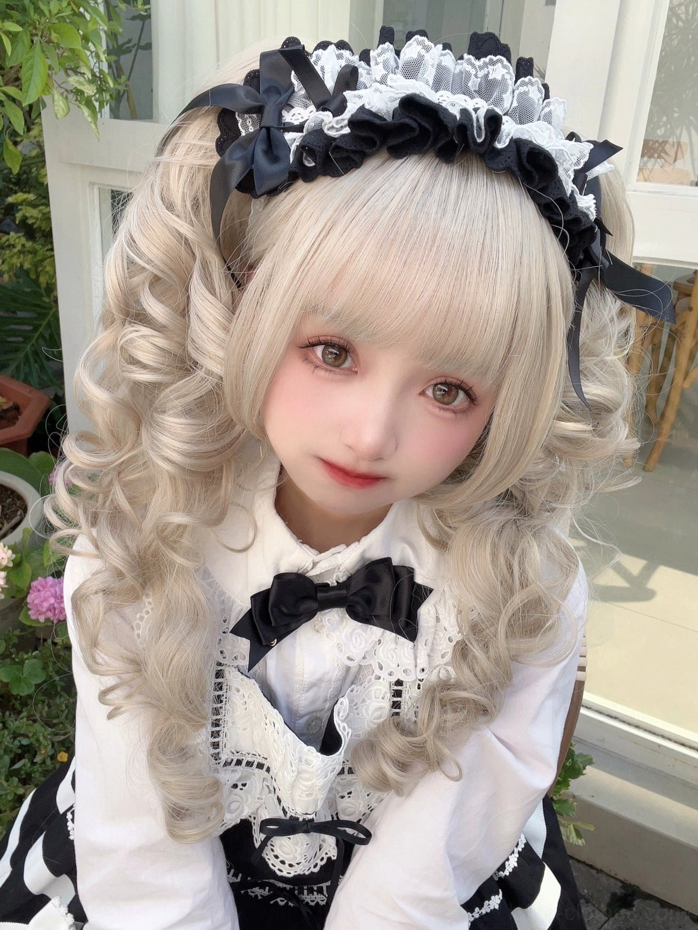 Black and white maid-style Lolita frilled lace headband with satin bow accents worn by blonde curly-haired model in outdoor balcony setting with green plants