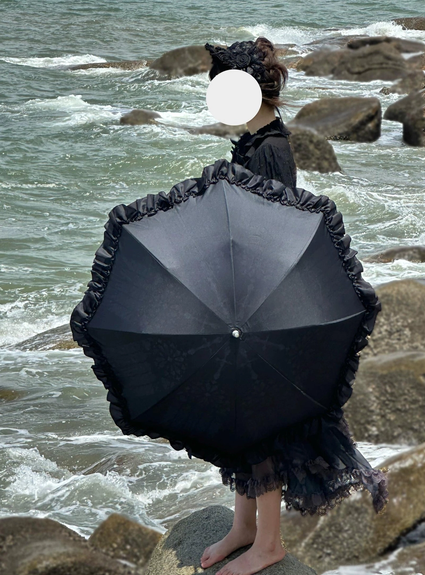 Model wearing black Lolita dress holding black ruffled parasol with window print pattern at scenic rocky seaside, back view demonstrating styling and practical use