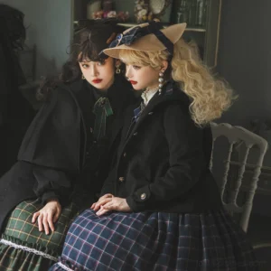 Two lolita models wearing matching black wool hooded coats and capes seated on vintage dining chairs indoors, displaying side-rear view with rounded cuff buttons and tailored silhouette