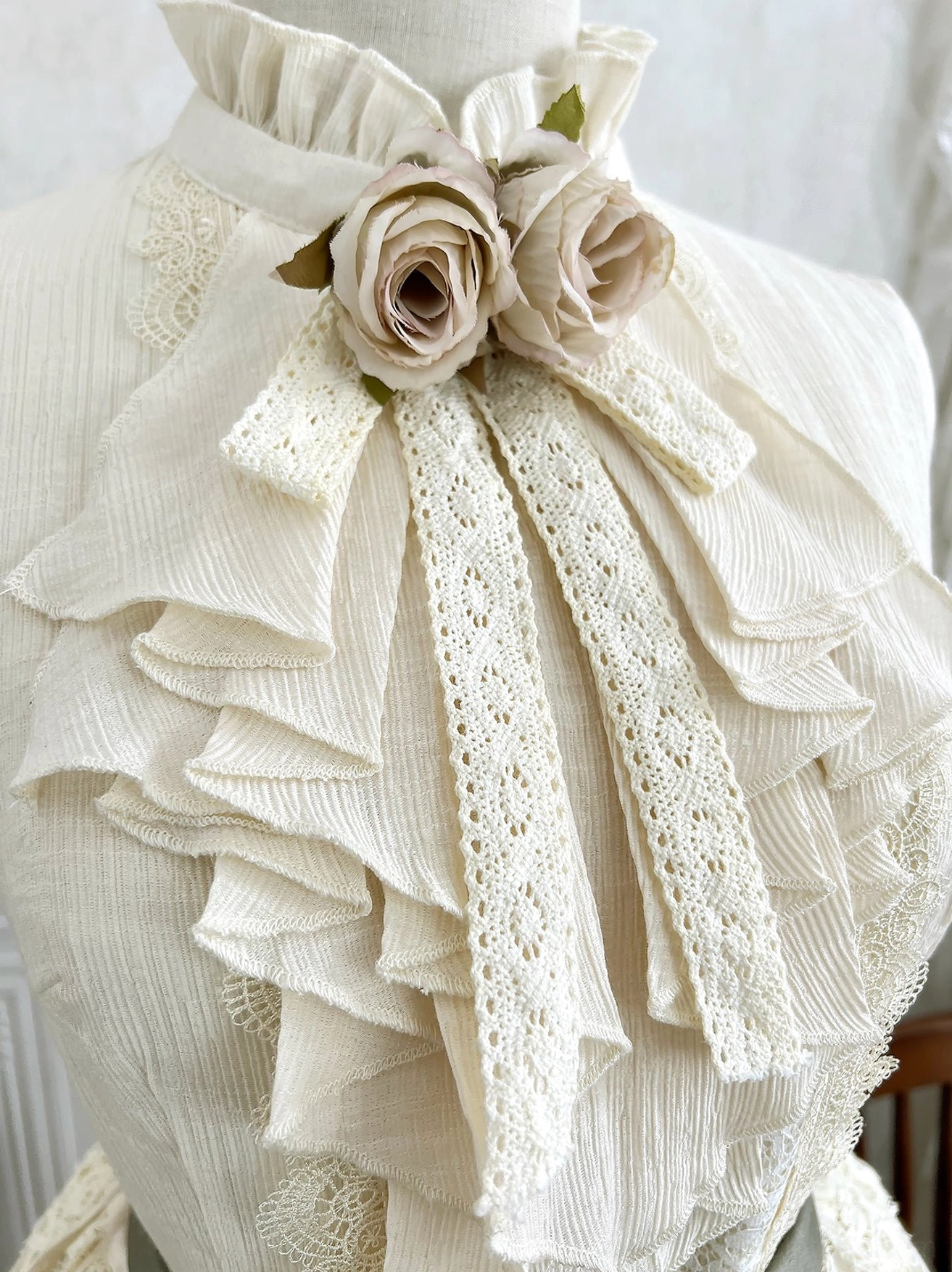 Close-up detail of sleeveless blouse collar and neck showing layered lace ruffles dual rose flower brooch and crepe fabric texture on dress form