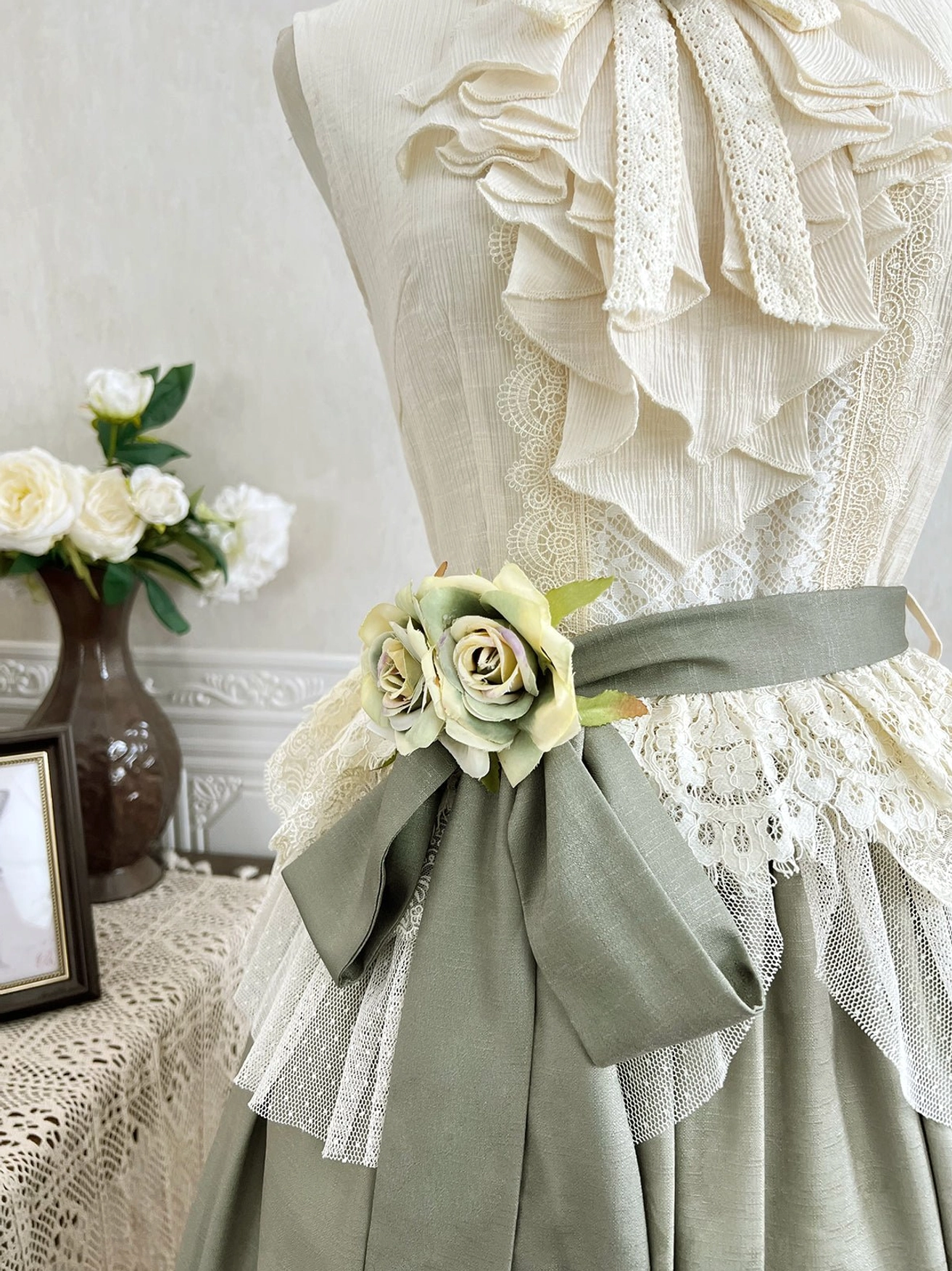 Close-up of blouse waistline showing scalloped cream lace trim and coordinating sage green waist sash with flower embellishment decorative detail
