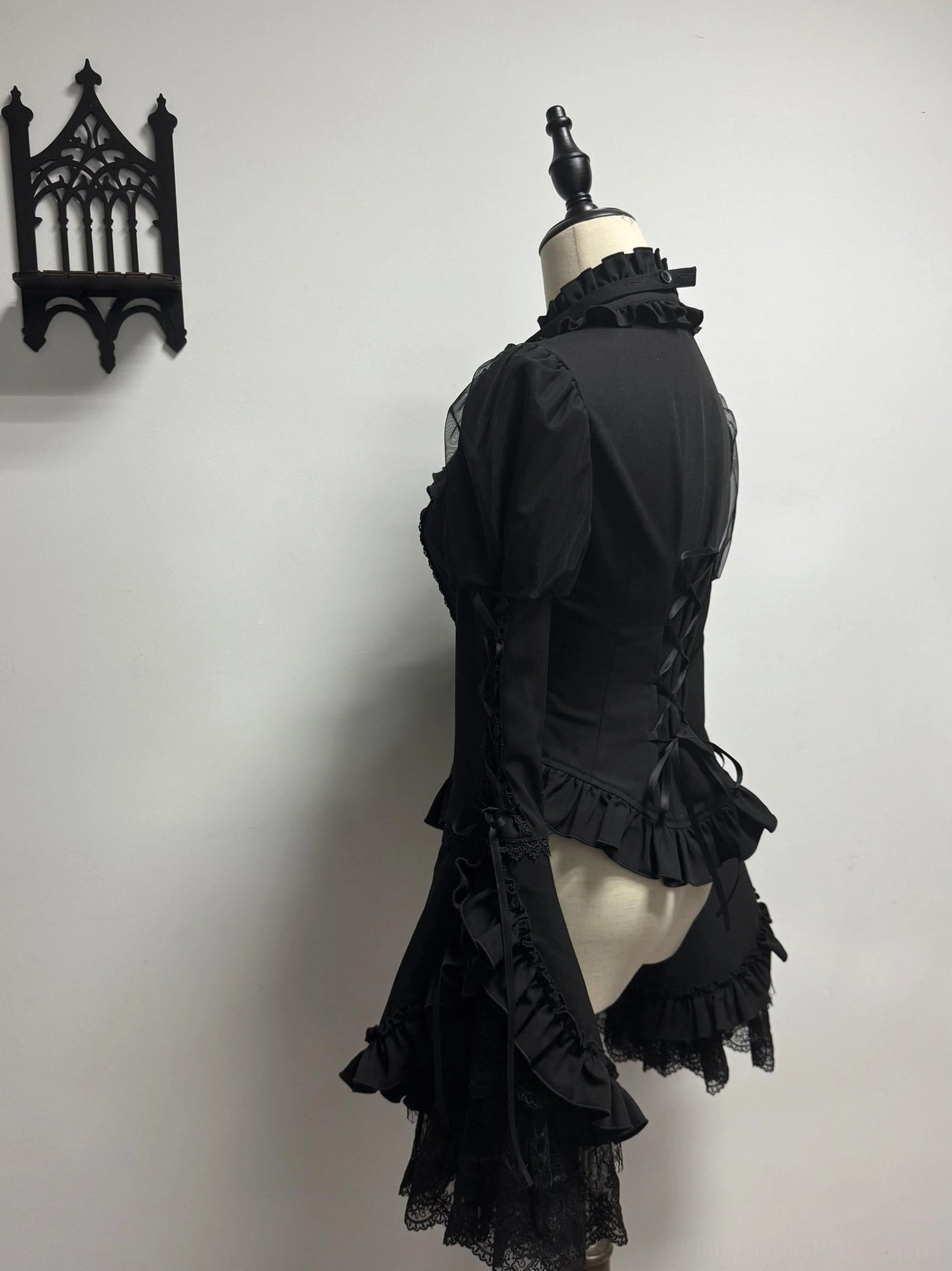Blue Blood Wedding Gothic Lolita corset blouse top side three-quarter view - asymmetrical ruffled waist hem, lace-up back design, solid black formal fabric, gothic wall ornament backdrop