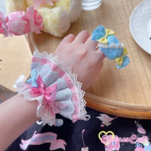 Close-up overhead tabletop shot of pastel blue gingham lace wrist cuff with pink bow detail and white scalloped lace edges displayed alongside candy-shaped hair clip on wooden dining table with plush toy and vintage tableware styling props
