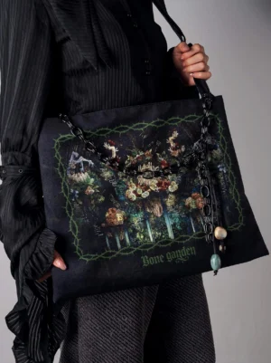 Bone Garden gothic tote bag front oblique view held by model, featuring black chain decorative strap with resin bead charms, paired with dark ruffled gothic blouse