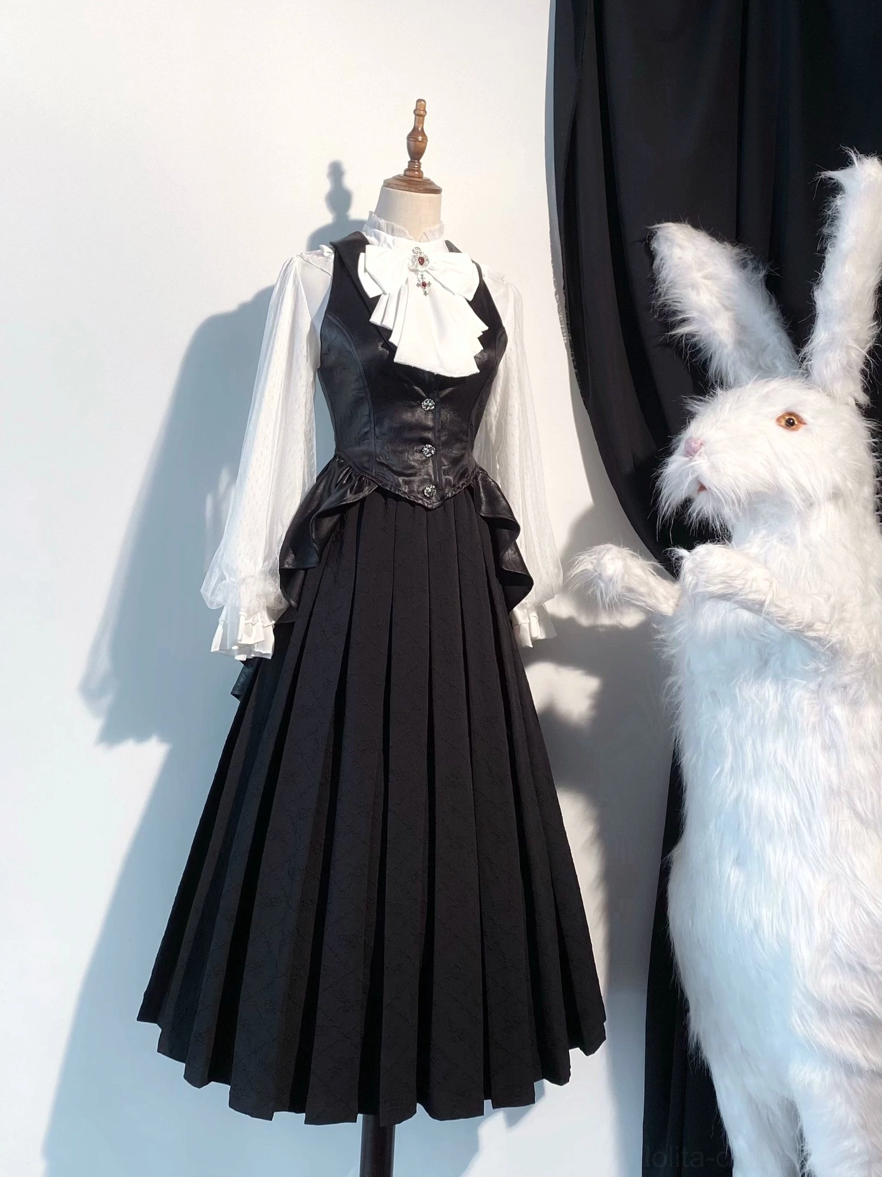 Front full-body view of black gothic lolita vest ensemble with white sheer mesh sleeves, long pleated skirt, white bow necktie with vintage brooch accent