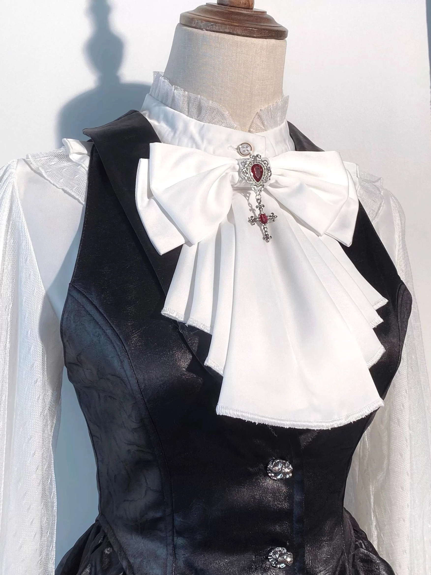Front close-up of gothic lolita waistcoat with white sheer ruffled stand-collar blouse, large white bow with gem-encrusted brooch, metal decorative buttons
