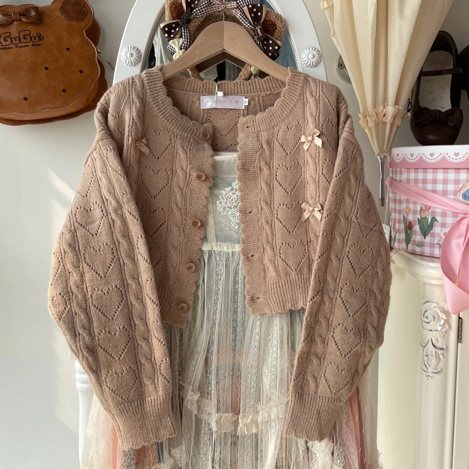 Brown cable knit hollow out Lolita cardigan with bow embellishments and round neckline displayed as flat shot, paired with sheer Lolita slip dress on vintage bedroom display