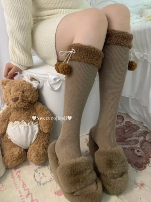 Brown over-the-knee Lolita fuzzy ball socks with lambswool pom-pom cuff and drawstring bow detail, worn with cream knit sweater and fluffy slippers in cozy bedroom setting