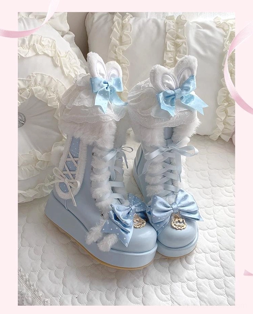 Overhead static flat lay of bunny-ear themed sky-blue lolita winter boots with 3D fluffy ear decorations, fur collar, lace-up design, polka-dot bows, and metallic toe ornaments on pillow backdrop