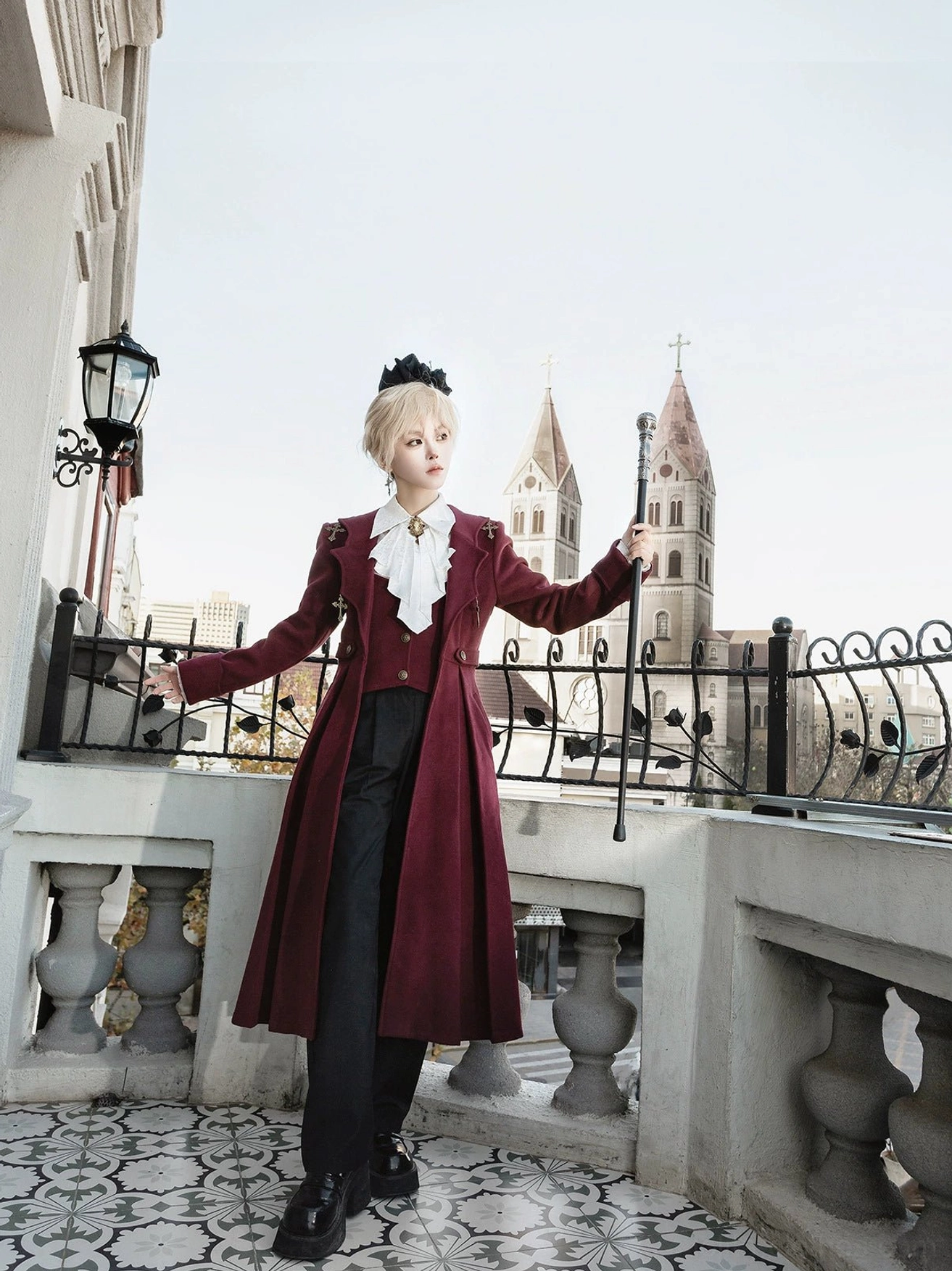 Gothic ouji lolita winter coat three-quarter side standing view model with walking cane burgundy velvet coat church spires background classic prince lolita aesthetic outdoor