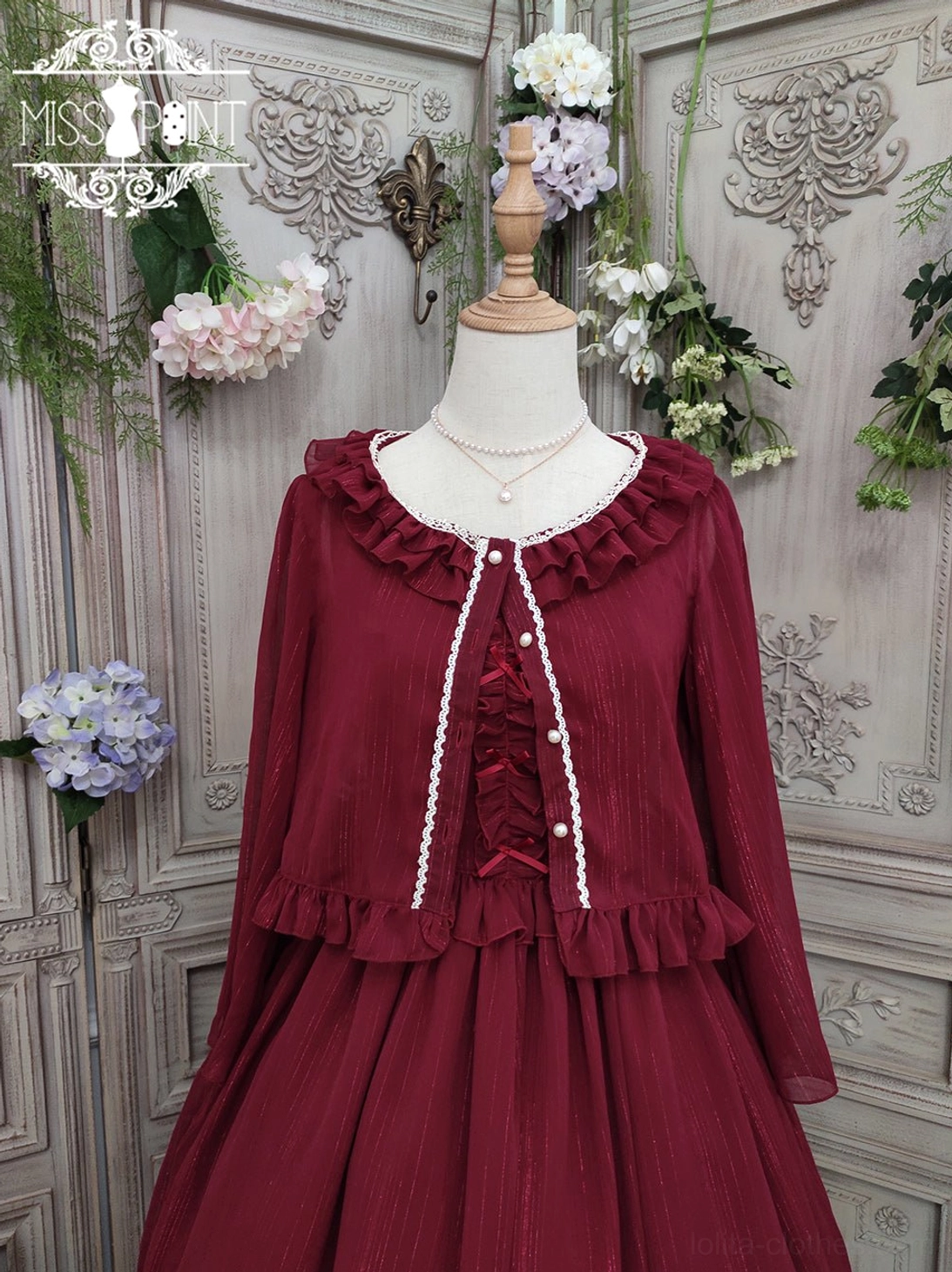 Burgundy lolita cardigan with ruffled crew neckline and shoulder ruffles, white trim lace-up front closure, ruffled waist trim, displayed in sheer chiffon fabric on dress mannequin, vintage floral garden setting