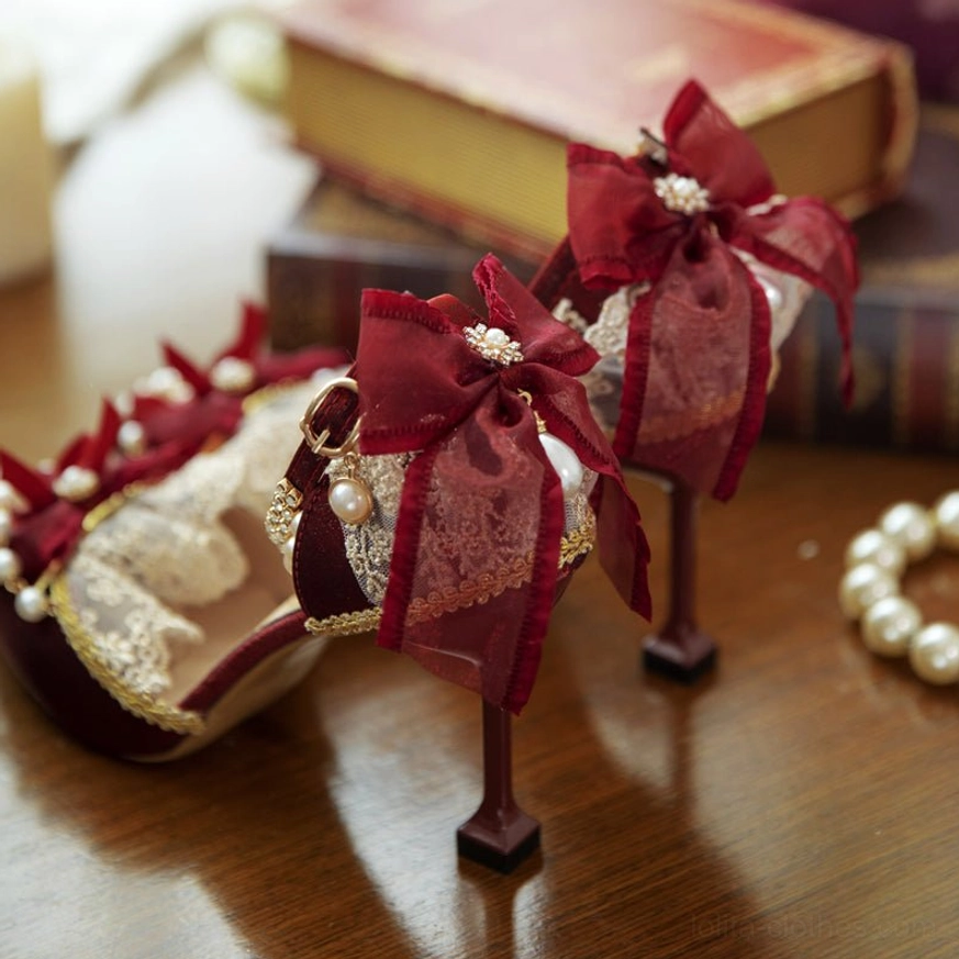 Burgundy pointed toe Lolita wedding heels three-quarter overhead view, featuring pearl-adorned red satin bow, intricate lace ruffles, gold trim edges, and elegant stiletto heel on wooden surface