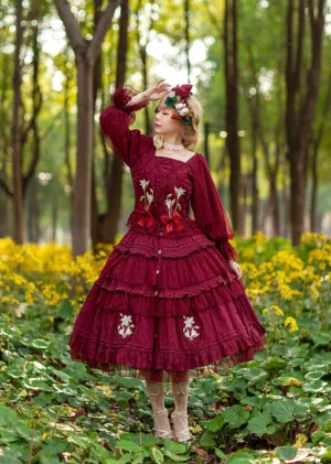 Burgundy red melaleuca lily embroidered country lolita dress featuring long bishop sleeves square neck tiered ruffled skirt with floral motifs worn by model in yellow flower garden