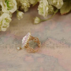 Close-up side flat lay view of cameo Lolita pearl ring with pink oval cameo portrait, delicate dangling pearl, filigree gold band on vintage patterned paper backdrop