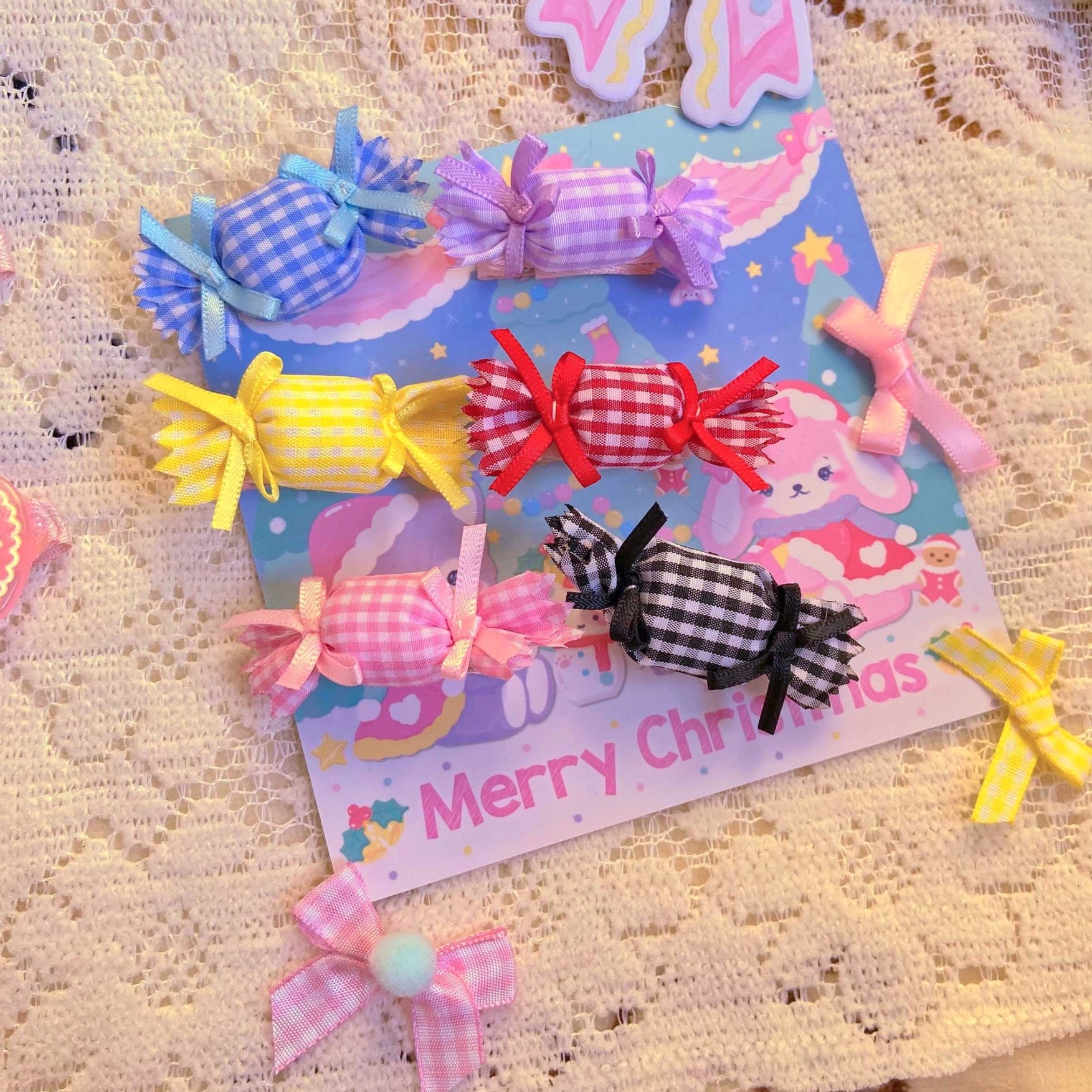 Assorted candy-shaped lolita hair clips and brooches in multiple gingham plaid colors displayed on lace tablecloth with Christmas greeting card, featuring pink yellow blue red purple black brooches and hair accessories with ribbon bows