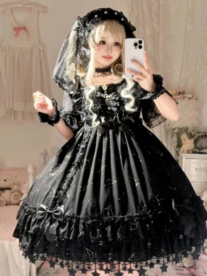Blonde lolita model wearing black constellation-themed OP dress with star hem tassels, matching veil and wrist cuffs in cozy bedroom setting