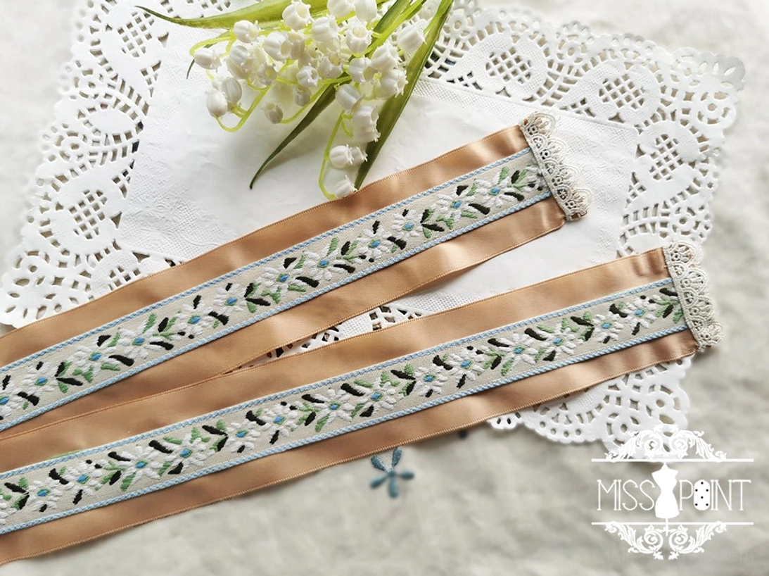 Top-down flat lay shot of tan champagne satin lolita ribbon belt with blue floral embroidery and pearl-trimmed lace ends on decorative doily