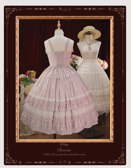 Cherry pink lolita JSK dress with matching half slip back view display showing lace-trimmed back corset adjustable spaghetti straps and layered ruffled skirt details in vintage indoor backdrop with dark red curtains and floral decorations