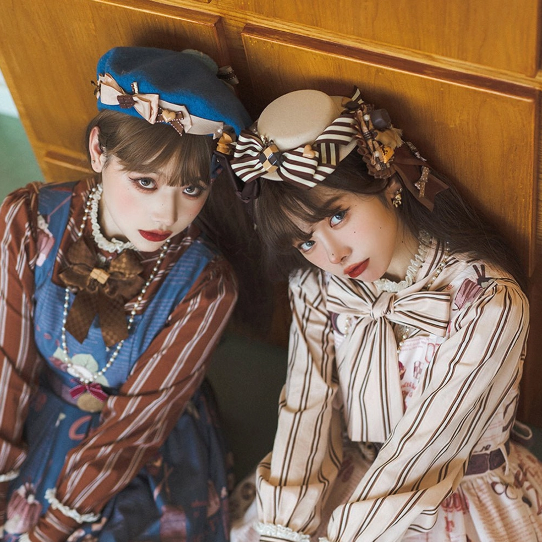 Two lolita models leaning on wooden door wearing chocolate-themed sweet lolita hats with striped bows and flat-top design, paired with chocolate print dresses in vintage home interior scene