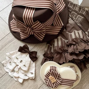 Classic chocolate lolita accessories flat lay display including brown bowler hat with striped ribbon bow, white embroidered lace jabot with heart details, and vintage striped decor on wooden desktop