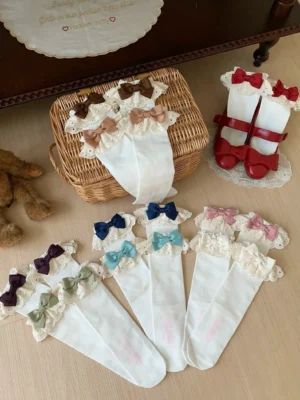 Overhead flat lay collection of assorted classic lolita calf socks with white base, double-layer ruffled lace cuffs, and multiple detachable bow colors including black, red, pink, blue, burgundy, navy, brown, and purple displayed on vintage dollhouse desktop with wicker basket and teddy bear decor