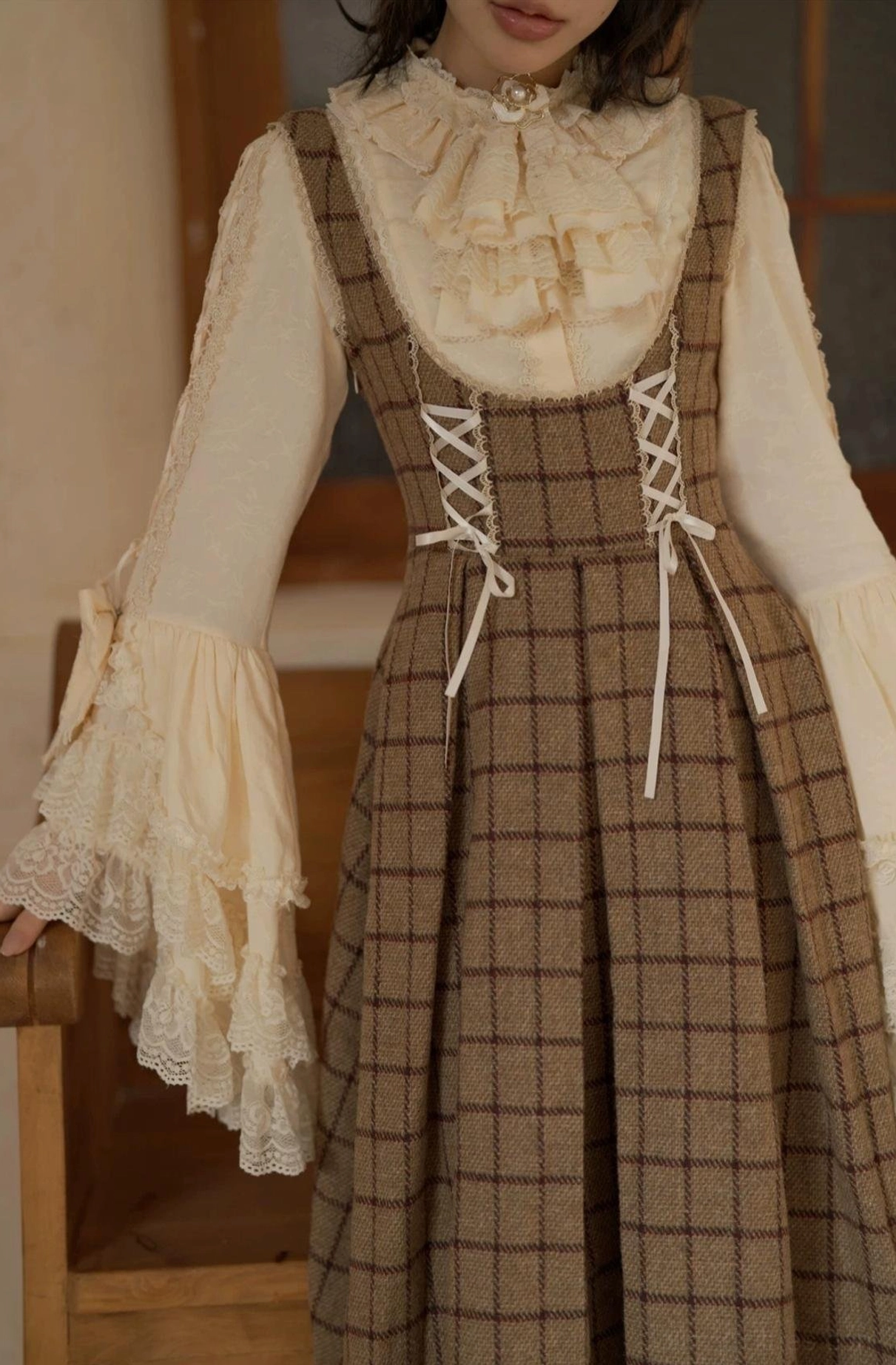 Coordinated lolita outfit featuring cream blouse with ruffled jabot and flared lace bell sleeves styled with brown tartan plaid overall dress with lace-up chest detail
