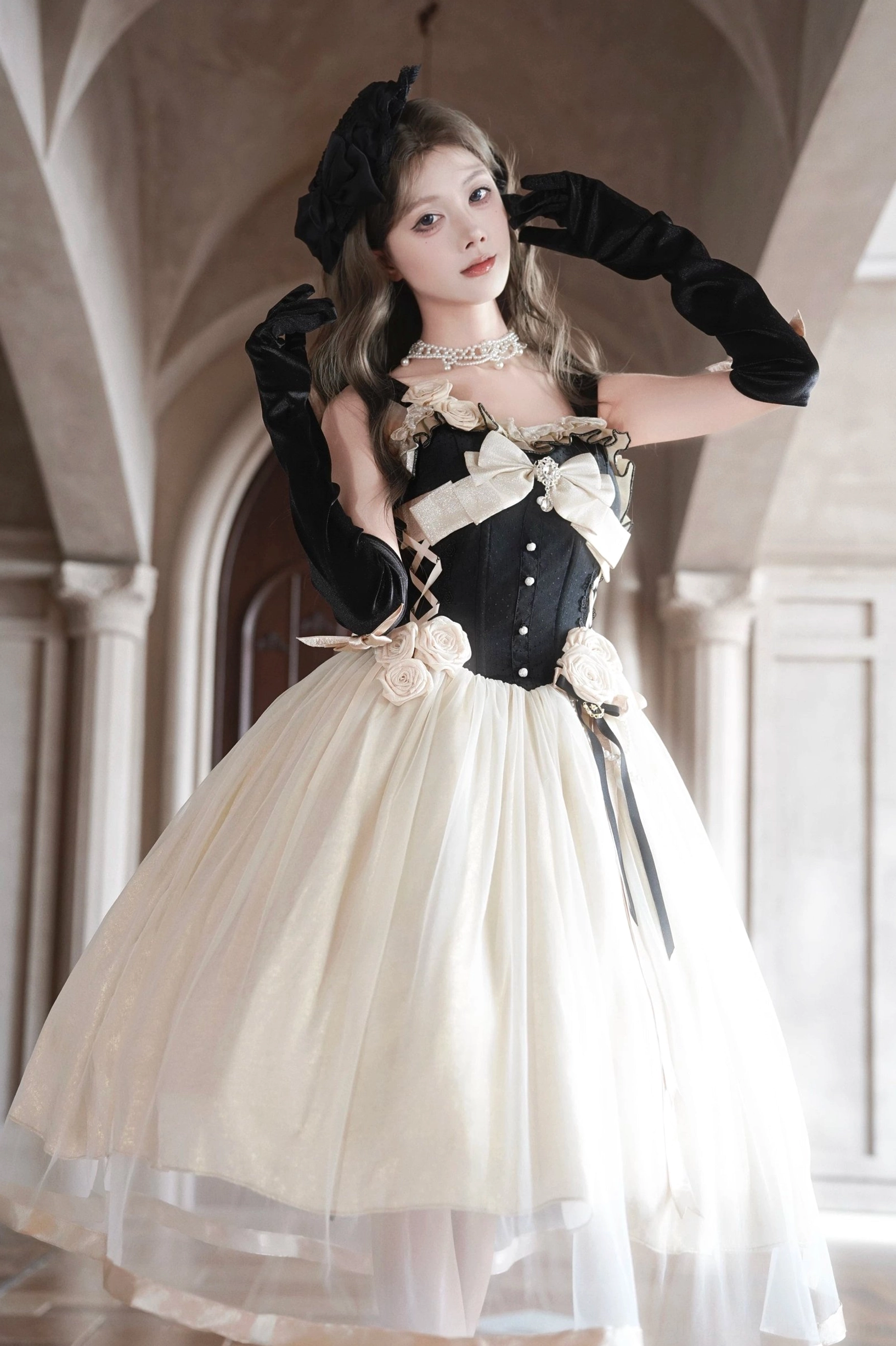 Low-angle front three-quarter view of Classic Lolita jumper skirt with model adjusting headwear, black satin gloves, large cream bust bow, 3D fabric waist roses detail