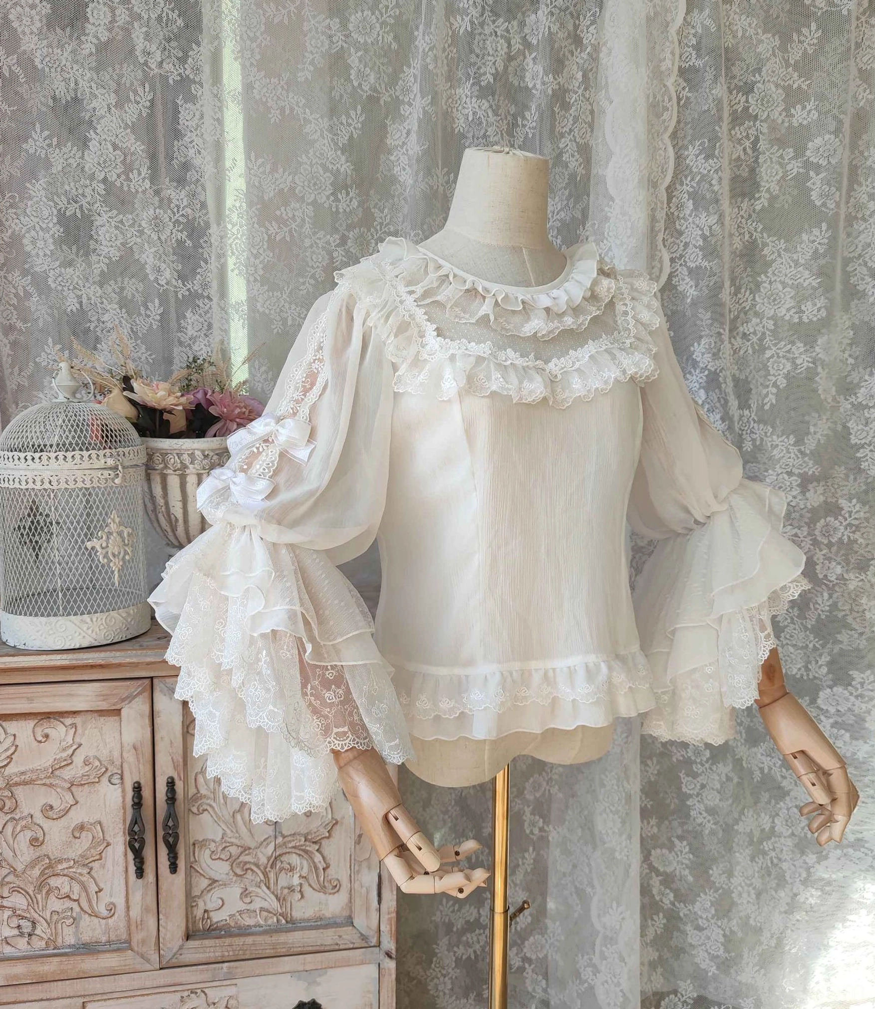 Off-white classic Lolita hime-sleeve blouse with ruffled round neckline and layered embroidered lace trumpet sleeves displayed on dress form with vintage lace curtain background