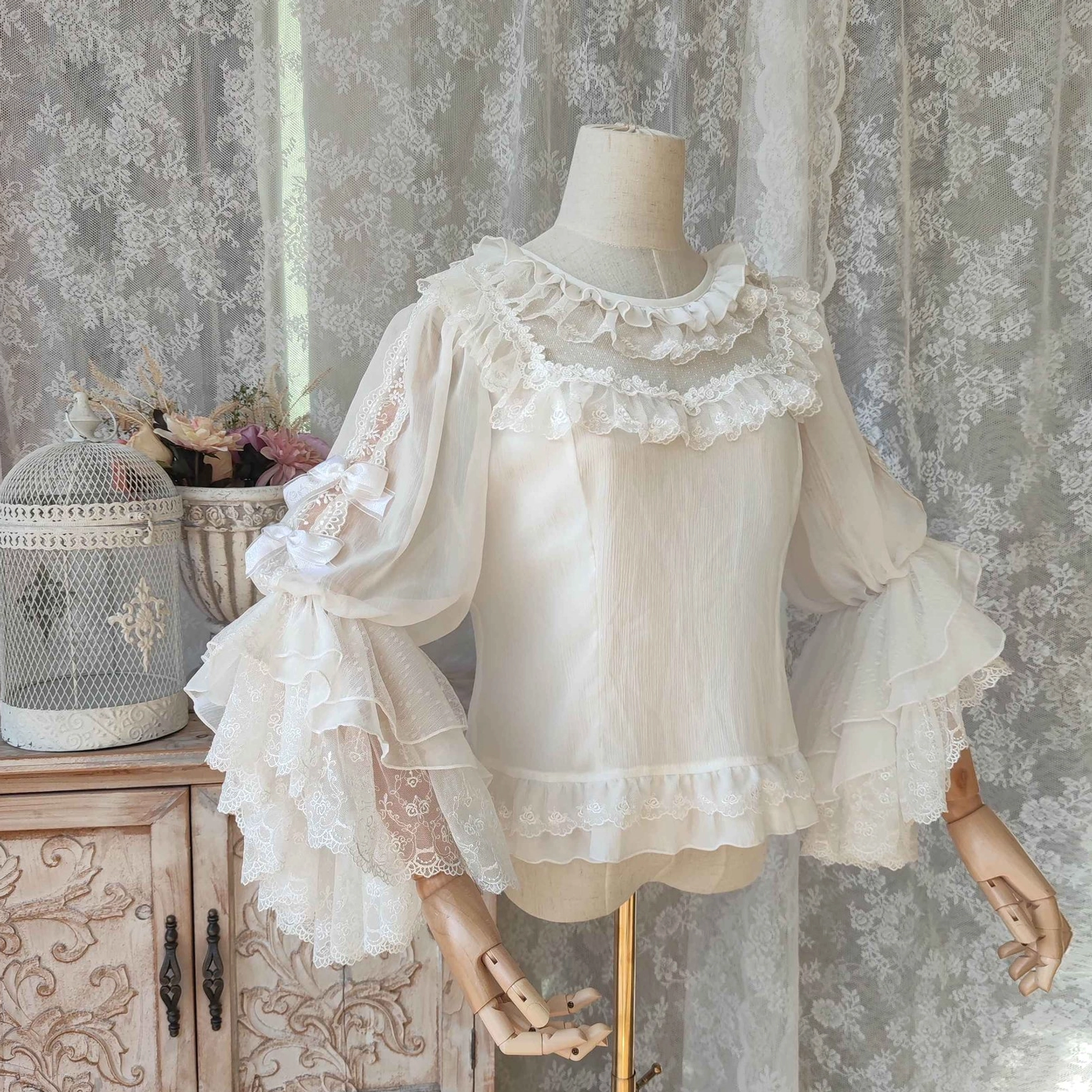 Off-white classic Lolita hime-sleeve blouse with puffed shoulders ruffled neck trim and layered lace sleeves with hem detail on dress form against vintage lace curtain background