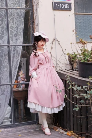 Plus-size model wearing pink classic Lolita OP dress with white detachable cuffs and ruffled collar, three-quarter rear view on residential balcony with lace window detail