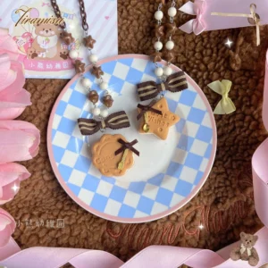 Top-down flat lay of kawaii cookie charm lolita necklace and hair clip set featuring brown striped bows, biscuit-shaped charms, and mixed beaded accessories on fluffy brown fabric with blue-white checkered plate and pink flower decoration