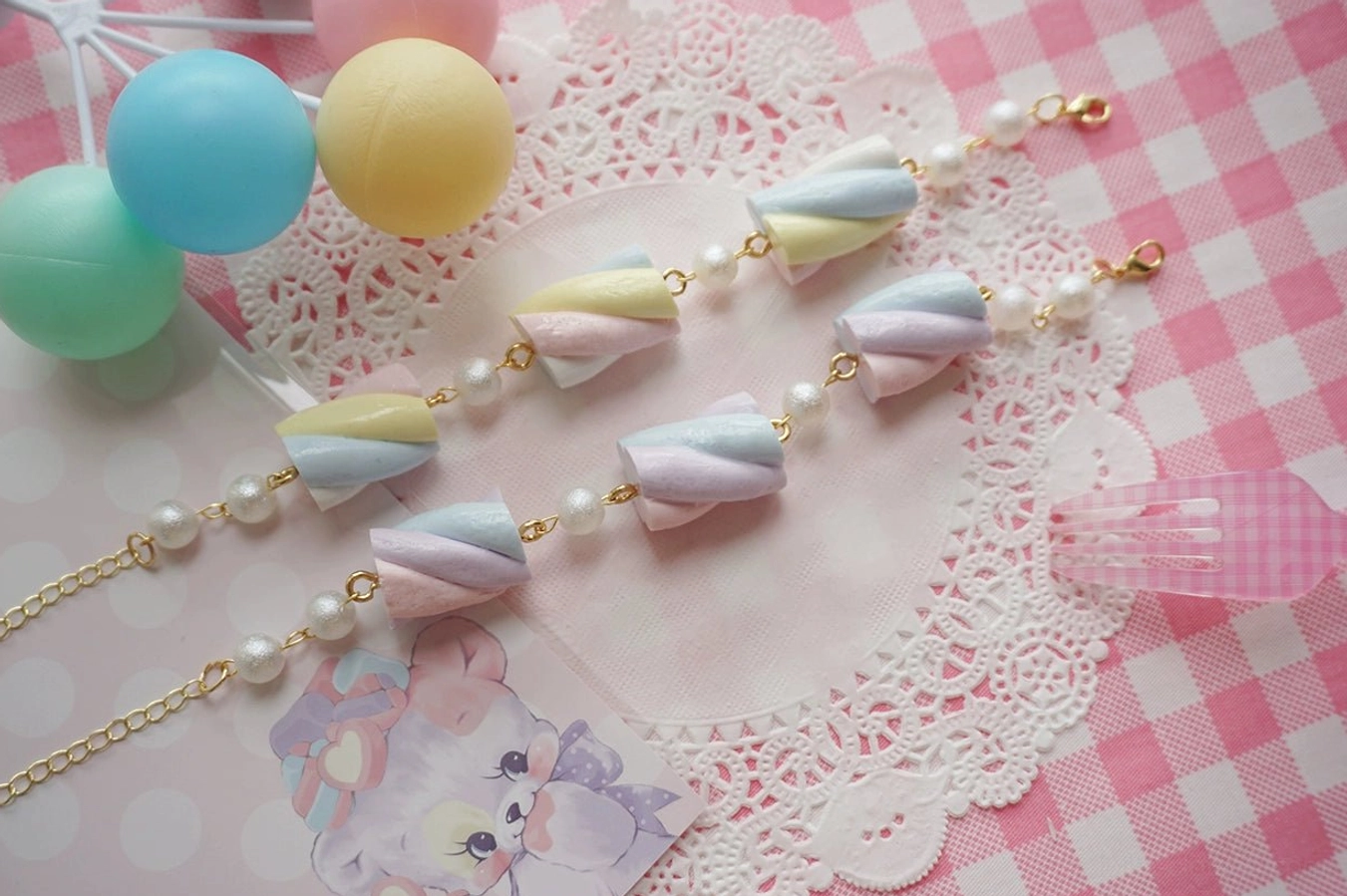 Top-down flat lay of pastel cotton candy resin lolita long necklace featuring multiple twisted marshmallow charms strung with imitation pearls, gold hardware, and adjustable extension chain on checkered tablecloth