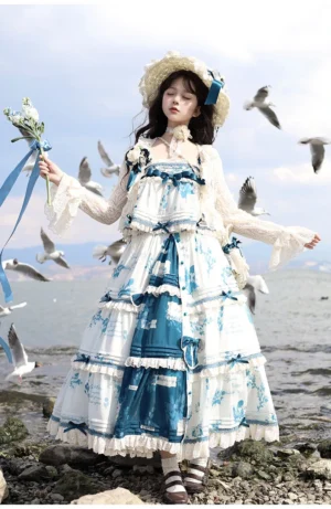 Front full-body view of blue and white porcelain printed Lolita slip dress set with detachable shoulder bows, tiered ruffled skirt, and coordinating straw hat with blue ribbon decor, photographed at seaside rocky beach with seagulls
