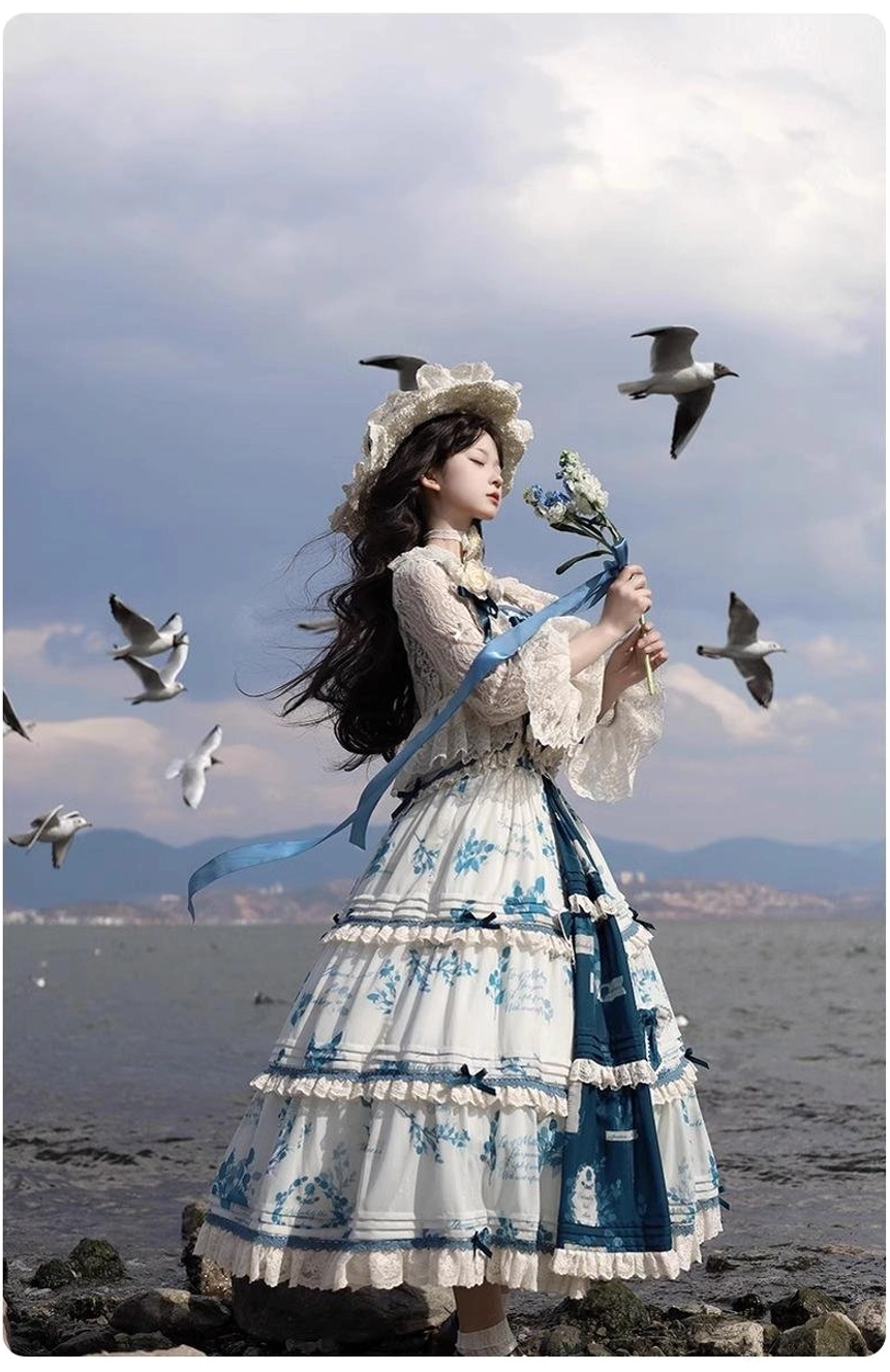 Side partial full-body view of Lolita dress with long lace sleeve cardigan set, flowing tiered skirt, blue waist ribbon, and lace-trimmed puff sleeves, photographed at seaside outdoor location