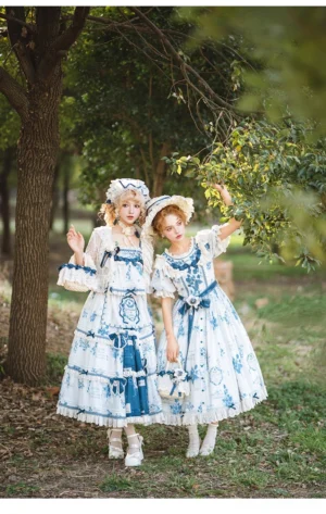 Two female models in coordinated blue and white printed Lolita dress sets with lace outfits, matching straw bonnets, and layered ruffled skirts, photographed together in woodland outdoor area