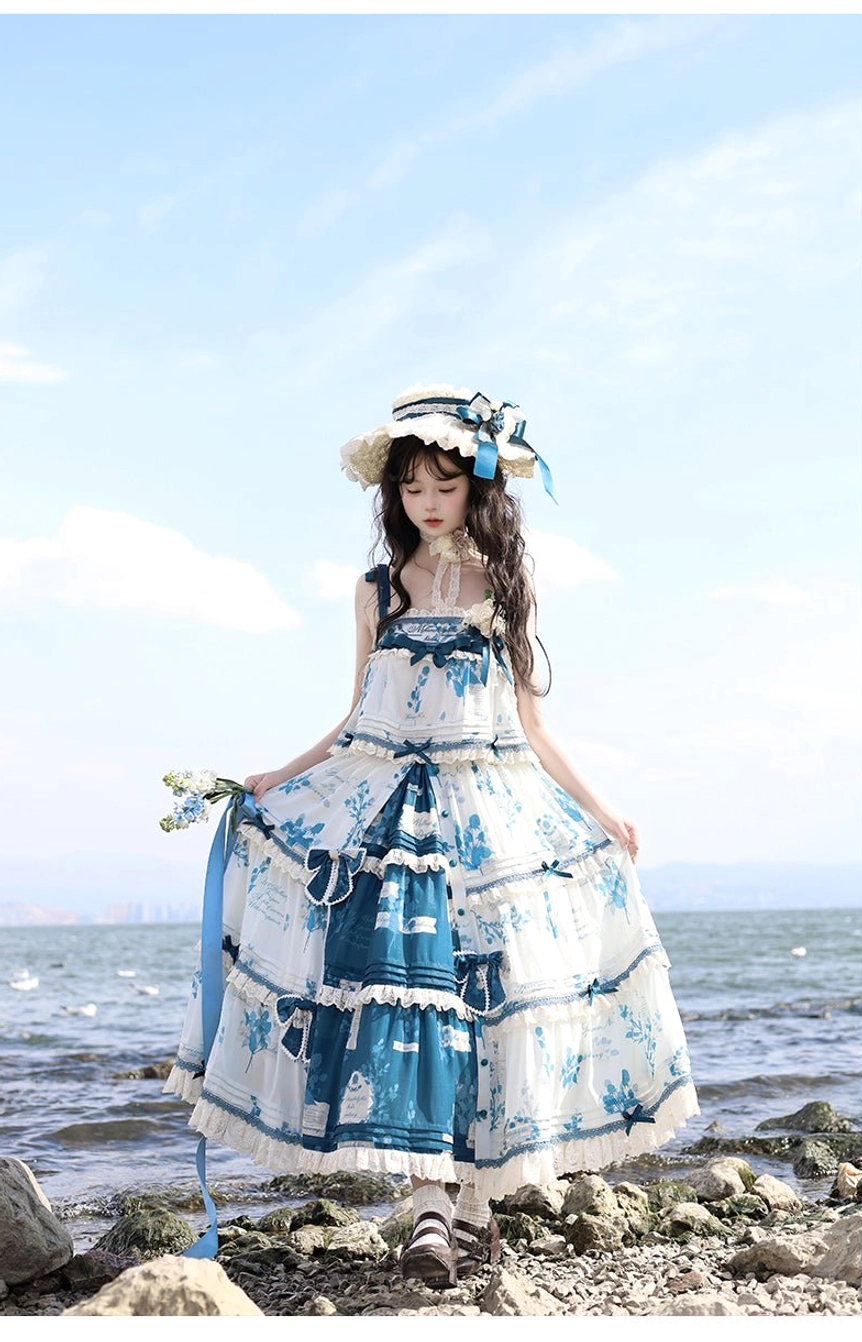 Three-quarter rear view of blue and white printed Lolita slip dress with square neckline, tiered skirt featuring blue ribbon sashes, and decorative straw bonnet, photographed at calm seaside rocky shore