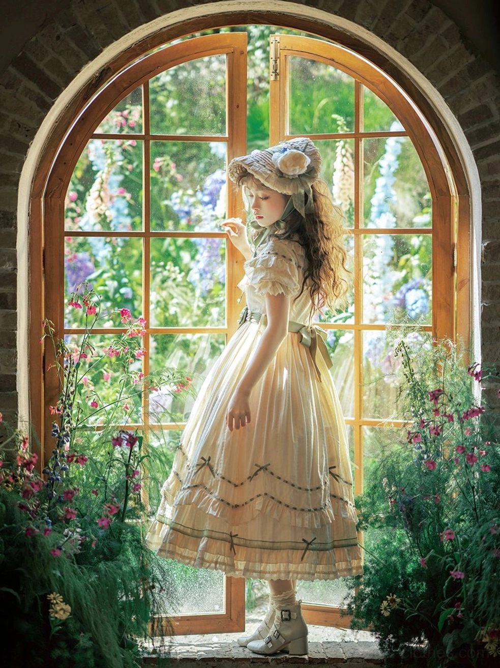 Three-quarter front view of lily of the valley country lolita dress with fitted bodice, lace trim, and tiered skirt with green ribbon accents by French window with garden blooms