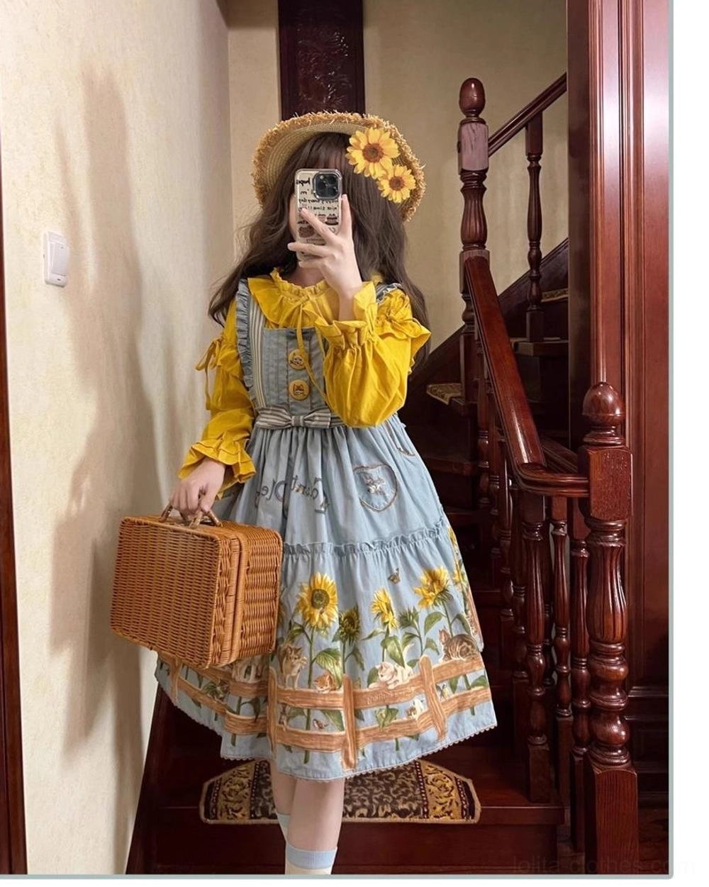 Three-quarter view of country lolita jumper skirt with sunflower cat print by wooden staircase, styled with woven picnic suitcase