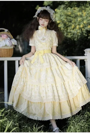 Three-quarter front full-body view of yellow cream country Lolita dress with high neck decorative bow, layered ruffled skirt with delicate floral embroidery prints, posed in grassy area beside white picket fence with decorative flower basket