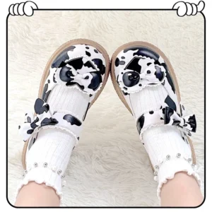 Cow print Lolita Mary Jane shoes with dual side bows displayed on fluffy white fur mat, showcasing glossy patent leather finish and thick round platform sole paired with frilly pearl-decorated white socks