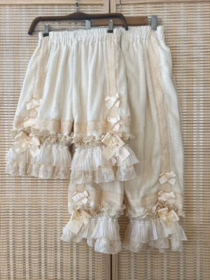 Cream-colored coral fleece lolita bloomers with tiered ruffled lace hems and decorative satin bows hanging on wooden display with bamboo woven vintage backdrop